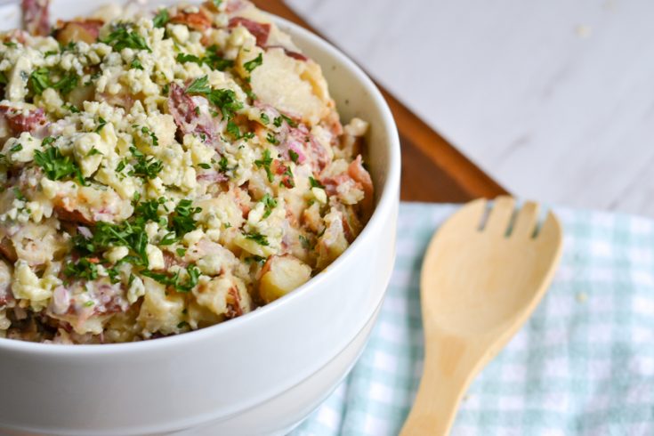 Bacon and Blue Cheese Potato salad in a white bowl next to a checked napkin and wooden spoon