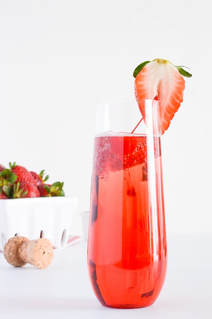 Pink bubbly drink in champagne flute, garnished with a cut strawberry and a strawberry basket in the background.