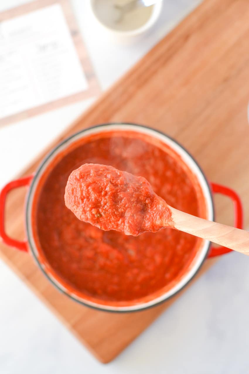 A wooden spoonful of homemade pizza sauce over a red sauce pan filled with sauce