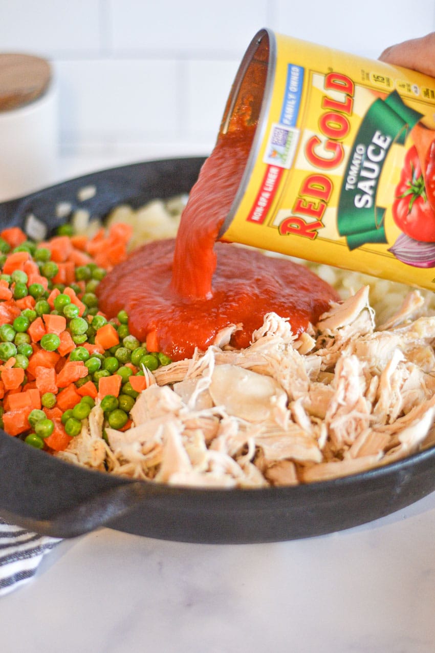 Black cast iron pan filled with onions, shredded chicken, and frozen peas and carrots, being topped with Red Gold Tomato Sauce