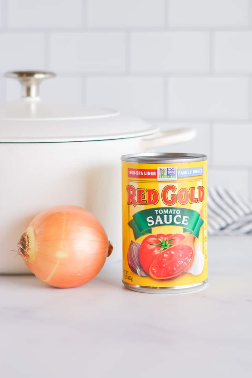 Can of Red Gold Tomato Sauce and yellow onion sitting in front of a large white dutch oven 