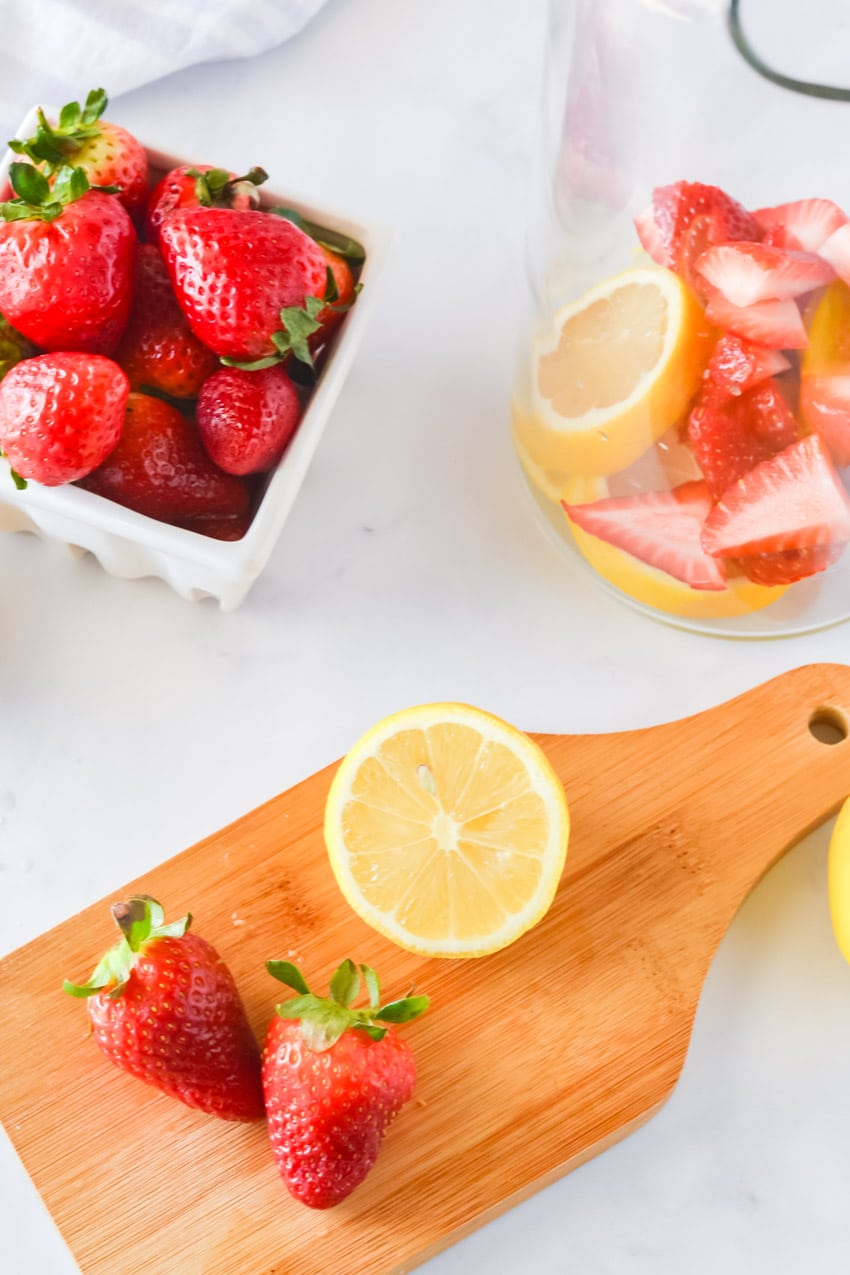 Strawberries and lemon on a small wood cutting board with a basket of berries and sliced fruit in a pitcher in the background 