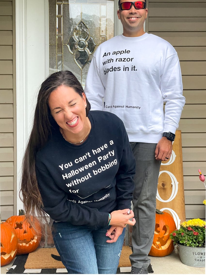 Couple standing on front porch that is decorated with jackolanters and mums, wearing a white and black sweater resembling Cards Against Humanity game