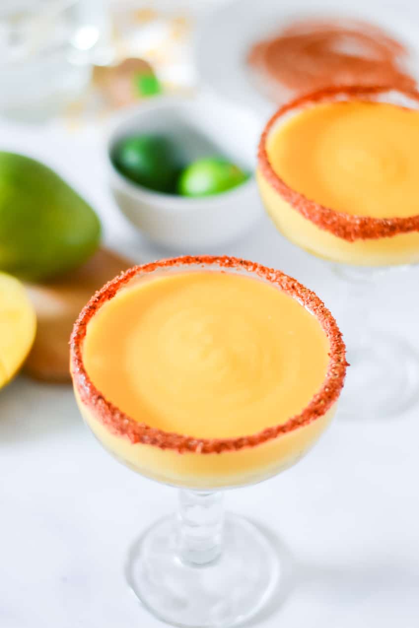 Two glasses of frozen mango margarita with fresh lime and mango to side on white background