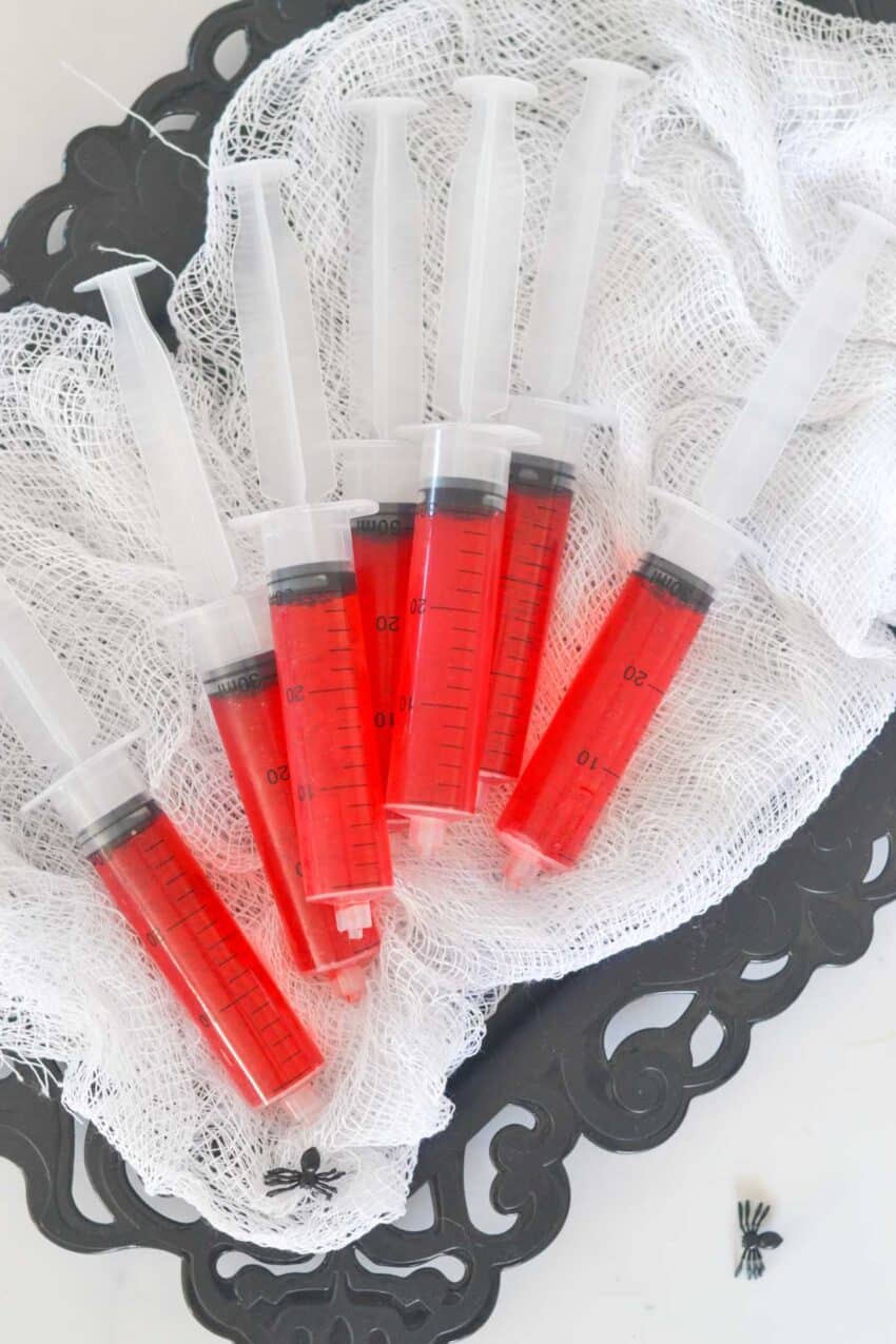 Several red jello shots in syringes laying on creepy white fabric on black tray.
