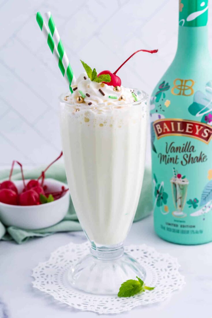 Bailey's milk shake topped with whipped cream and a cherry in tall glass with a bottle of Bailey's and a small bowl of cherries in the background.