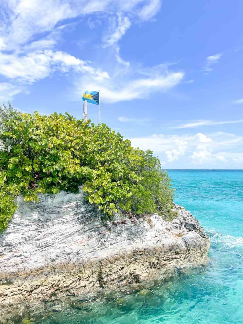 A rocky jetty in the Bahamas with vegetation growning on top, and a Bahamian flag with the blue ocean and sky in the background.
