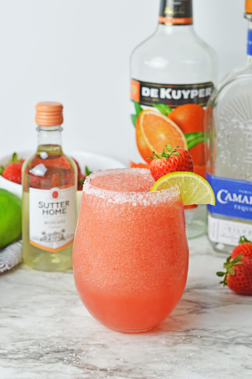Glass of Strawberry Moscato Margarita with ingredients in the back: limes, white bowl filled with strawberries, moscato wine, triple sec, and tequila.