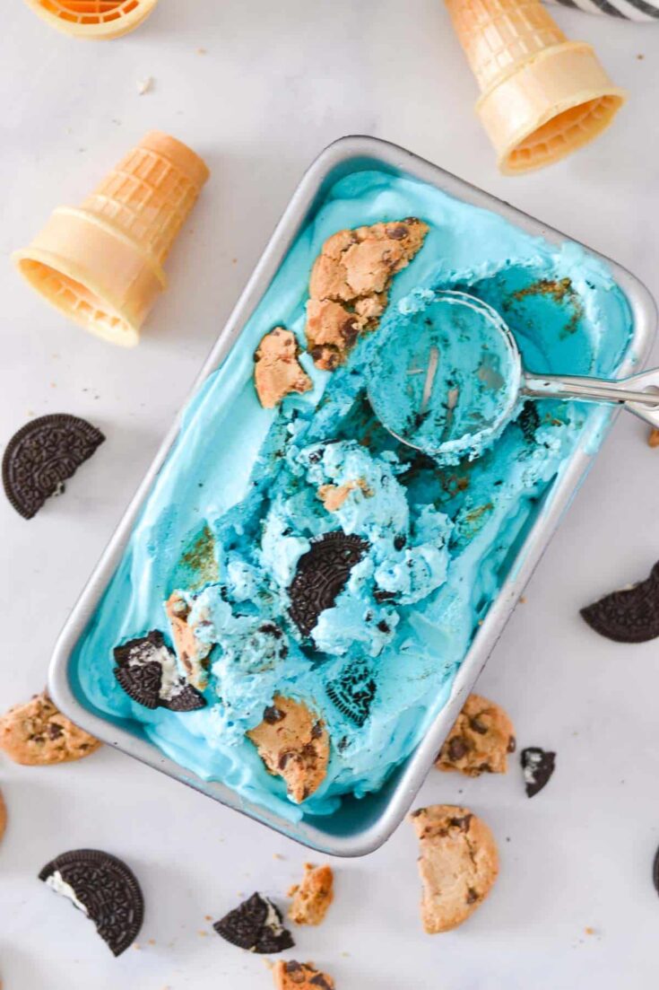 Loaf pan filled with blue ice cream containing chunks of Oreo and chocolate chip cookies, with cookie pieces sprinkled around.