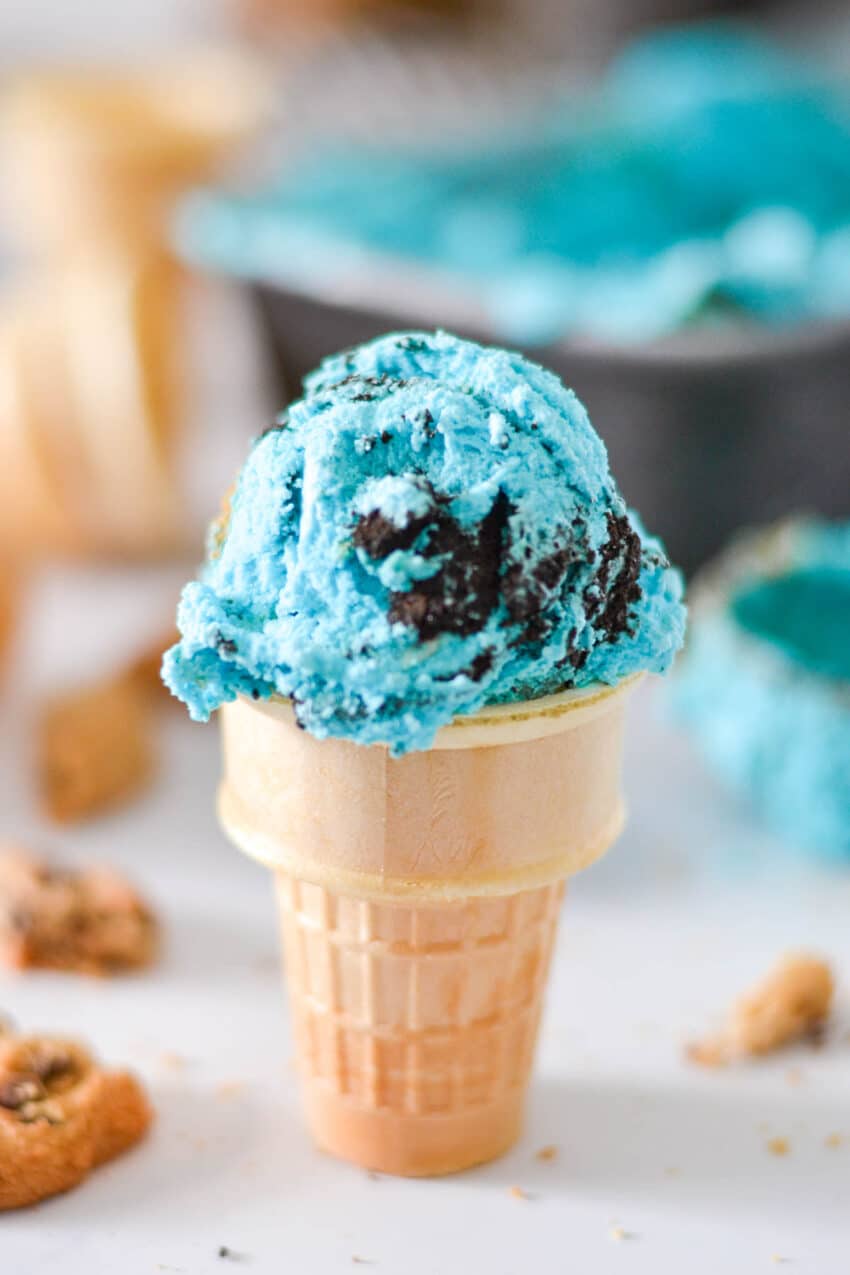 Scoop of blue cookie monster ice cream on cake cone, with more ingredients in the background