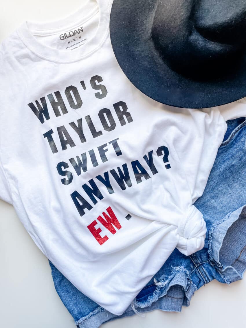 White t-shirt that says "Who's Taylor Swift Anyway?" in black text, and "EW" in red text. Style with a black fedora and jean shorts. 