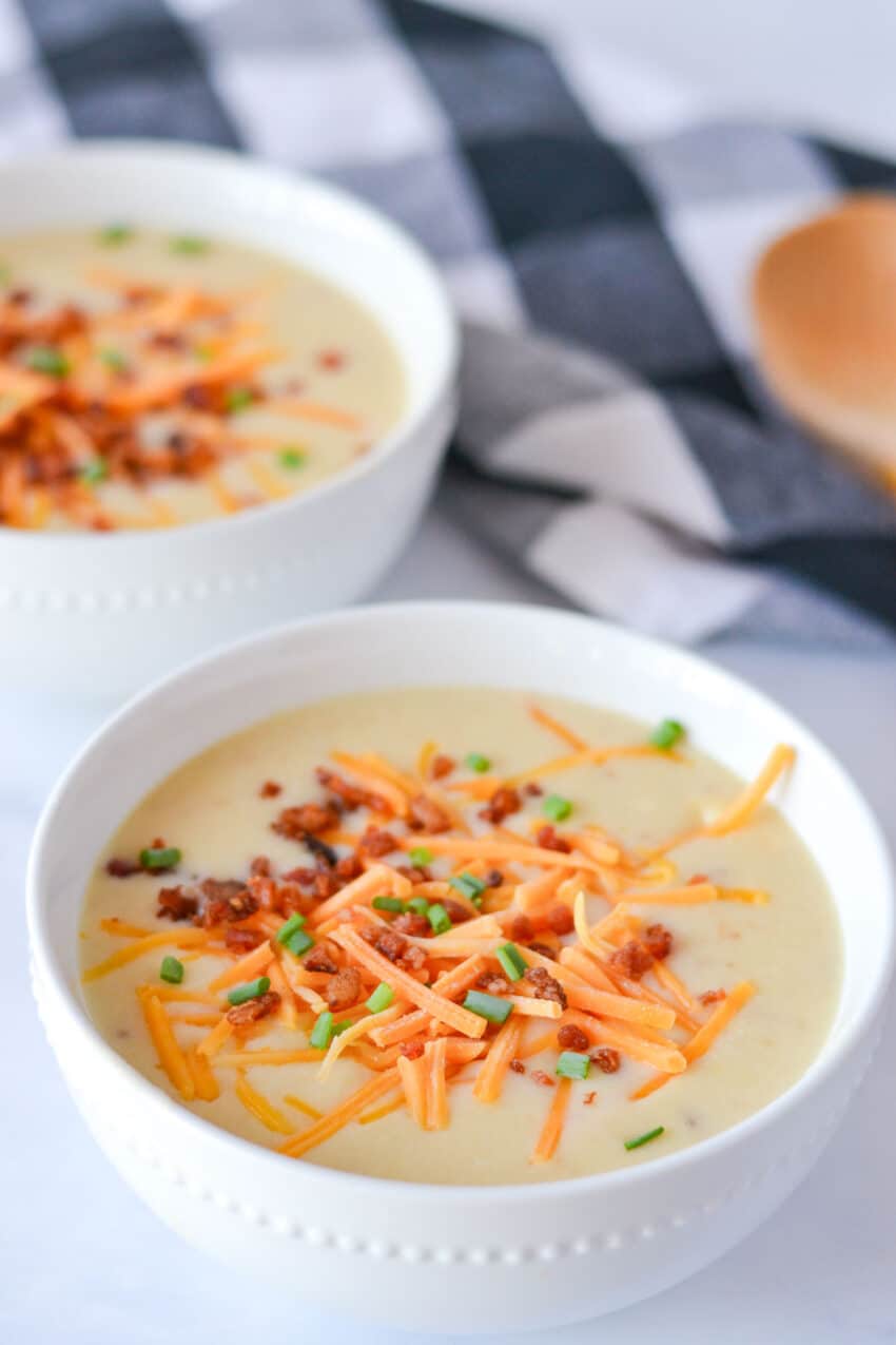 Close up of a white bowl filled with potato soup and topped with bacon bits, shredded cheddar cheese, and green onions