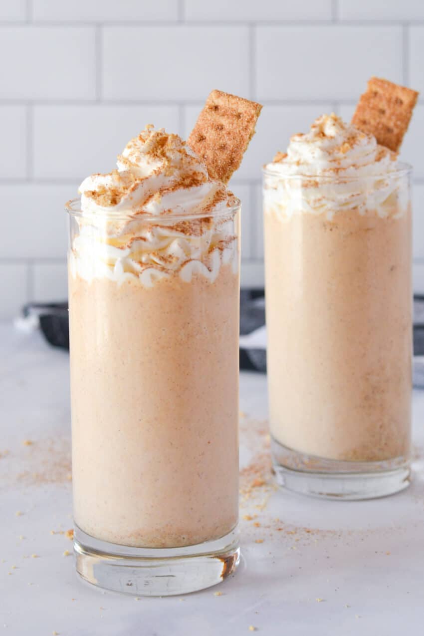 Two glasses of pumpkin pie milkshake topped with whipped cream and garnished with a sprinkle of graham cracker and pumpkin spice