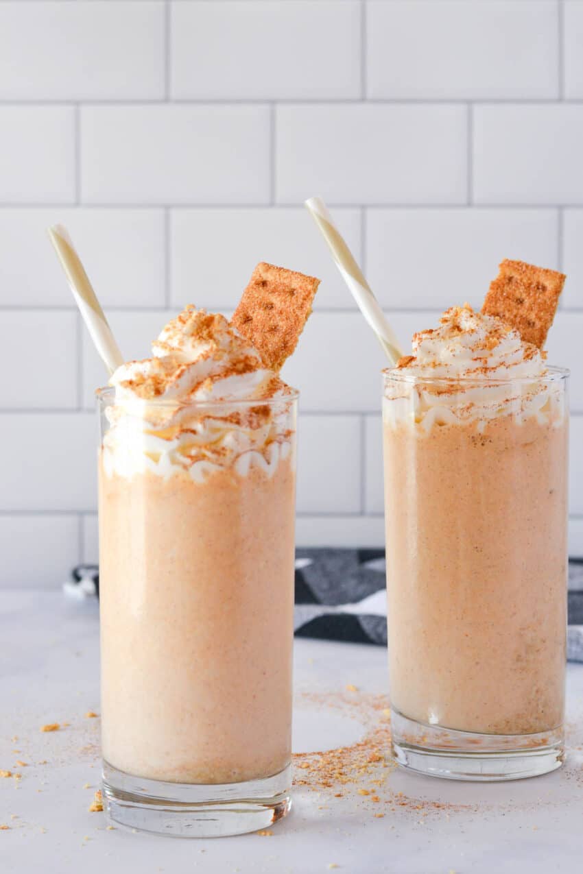 Two tall skinny glasses filled with pumpkin pie milkshake, and topped with a large swirl of whipped cream, and a dusting of graham cracker and pumpkin pie spice