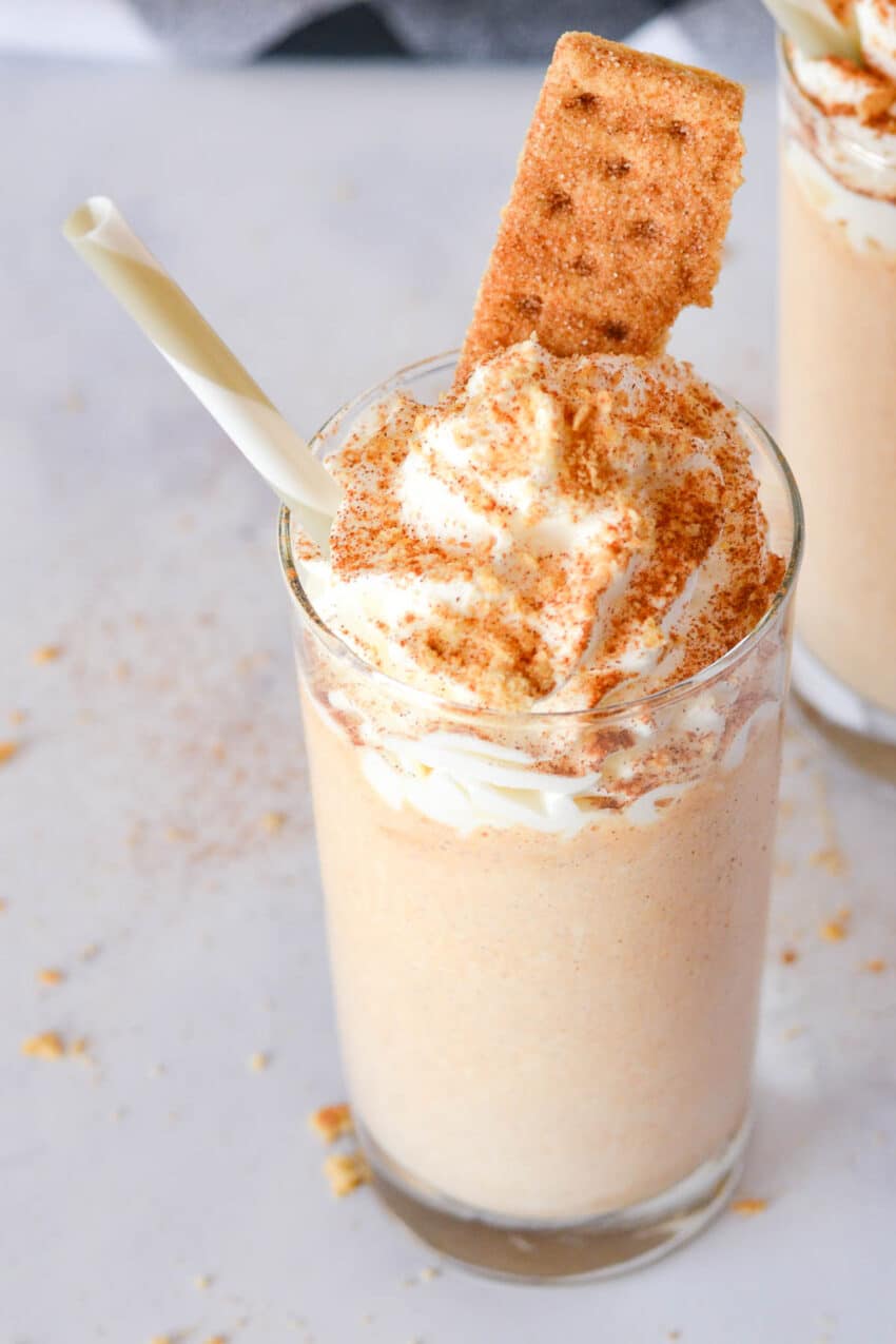Glass of pumpkin spice milkshake with whipped cream and graham cracker on top, with a straw.