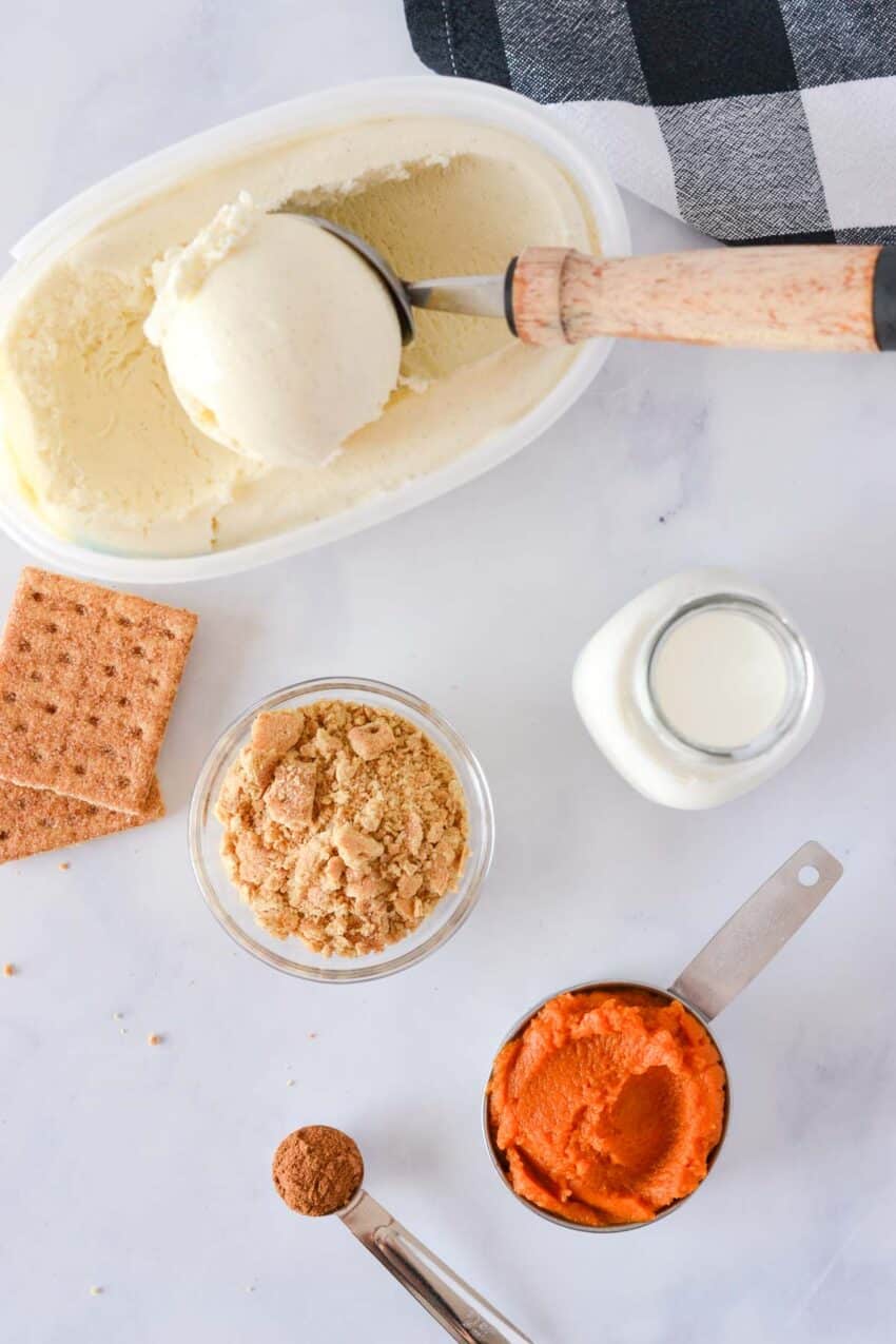 Ingredients for a pumpkin pie milkshake: a tub of vanilla ice cream,  crushed and whole graham crackers, a glass jar of milk, a measuring cup filled with pumpkin puree, and a teaspoon filled with pumpkin pie spice. 