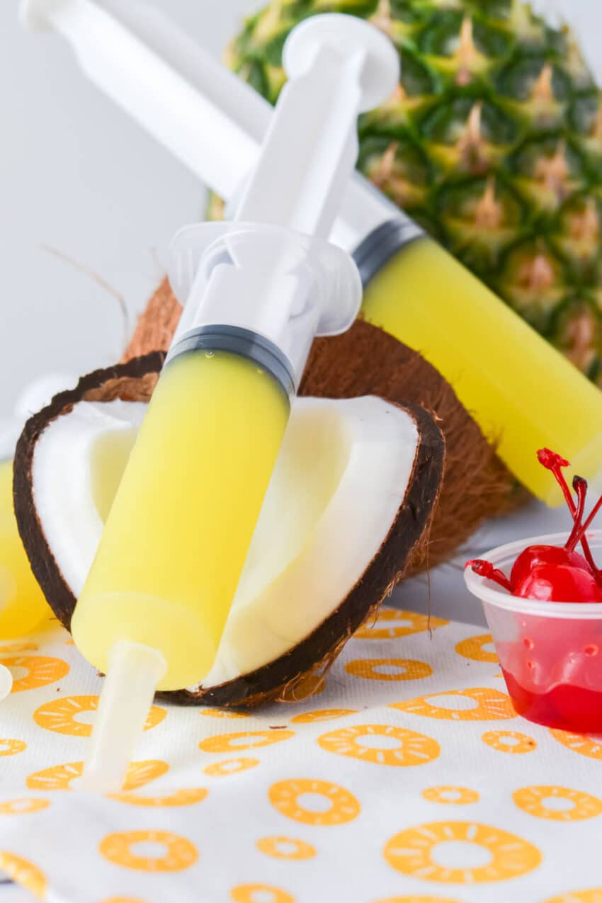 Yellow Piña Colada Jello Shots in a plastic syringe balanced on coconut shell with cherries and pineapple in the background.